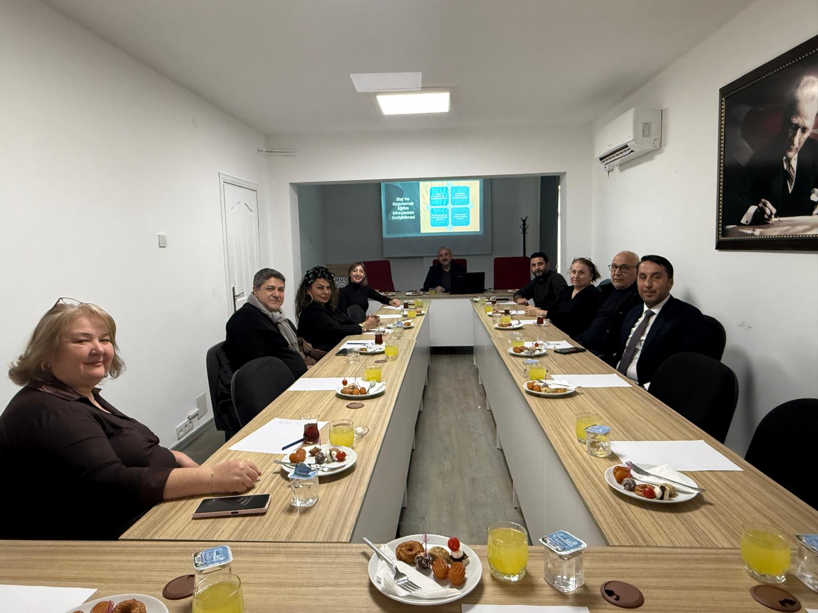U bir masa etrafında kadın ve erkeklerden oluşan yaklaşık 10 kişi gülümseyerek kadraja girmiş. 